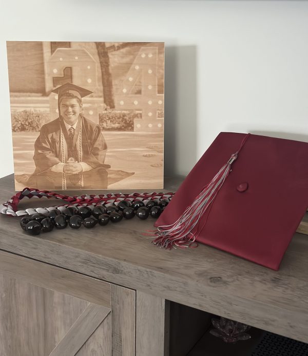 A maroon graduation cap, tassel, leis, and an engraved photo of a test school graduate are displayed on a wooden cabinet.