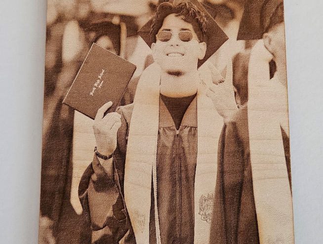 A graduate in cap and gown smiles, holding a diploma with a peace sign at a graduation ceremony, celebrating achievements that could be commemorated with personalized gifts from Laser Engraving Services.