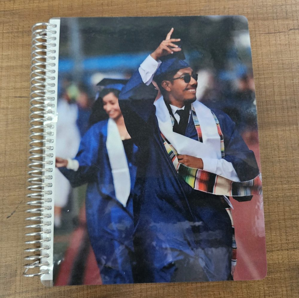 A graduate in a blue cap and gown celebrates, smiling and gesturing, during a ceremony on a spiral notebook cover—perfect for personalization with Laser Engraving Services.