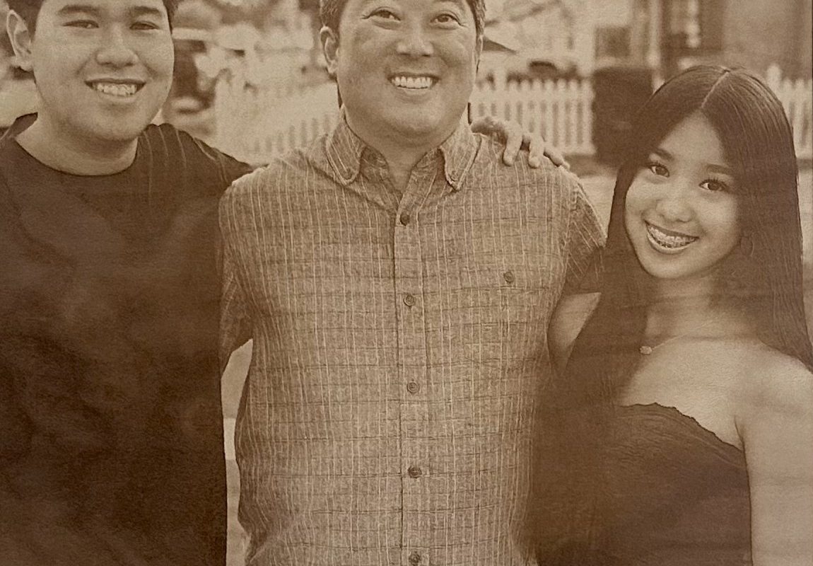 Sepia-toned photo of three people smiling, standing close together outdoors in front of a white fence—perfect for transforming into a cherished keepsake with our Laser Engraving Services.