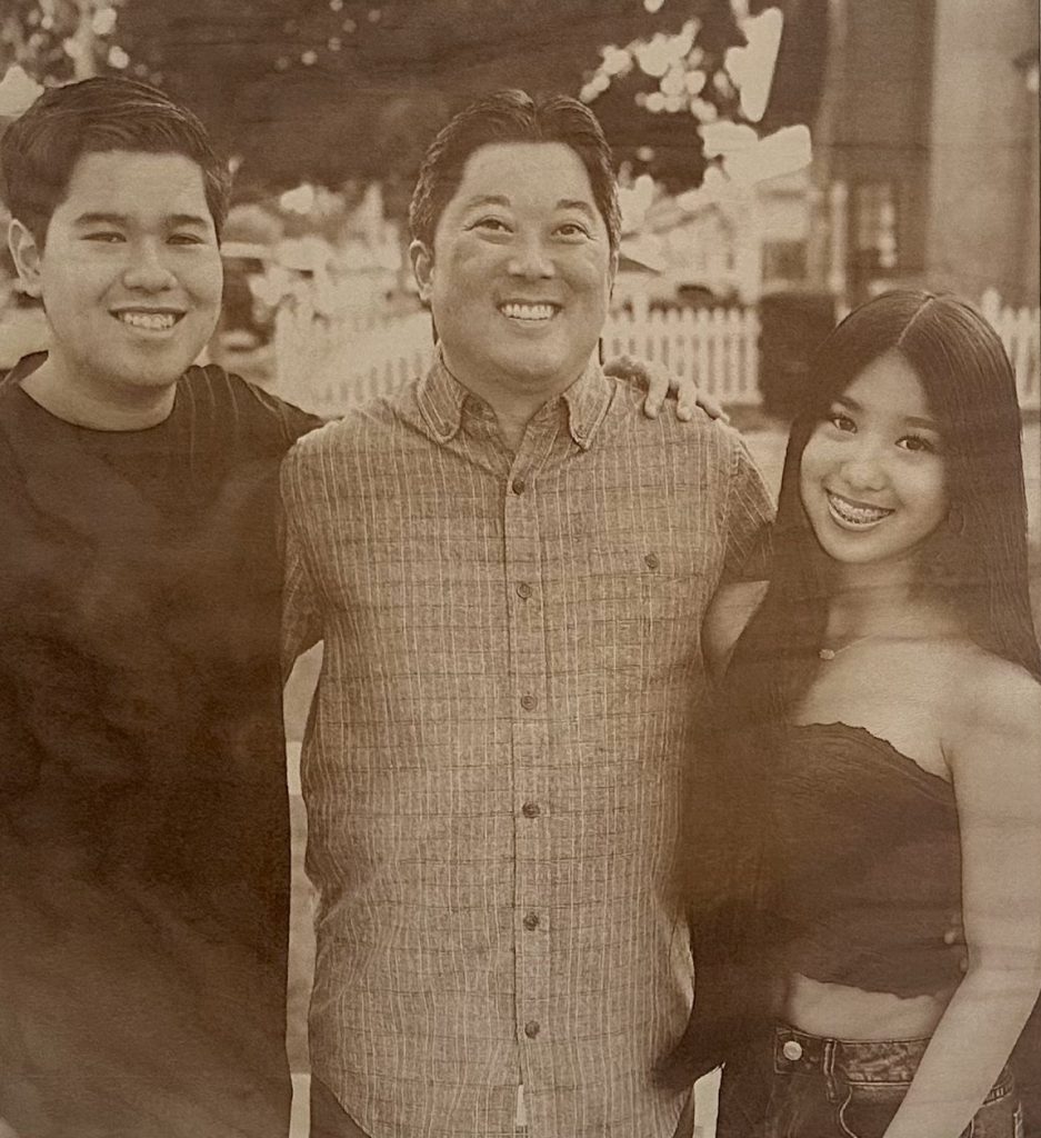 Sepia-toned photo of three people smiling, standing close together outdoors in front of a white fence—perfect for transforming into a cherished keepsake with our Laser Engraving Services.