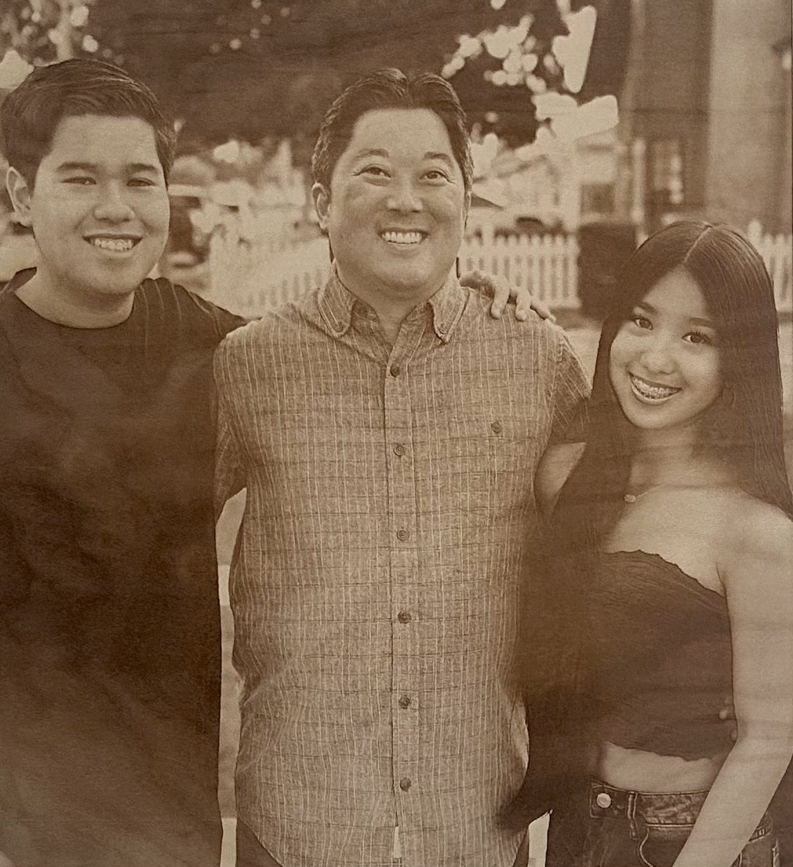 Reannas Fam Sepia-toned photo of three people smiling, standing close together outdoors in front of a white fence—perfect for transforming into a cherished keepsake with our Laser Engraving Services.