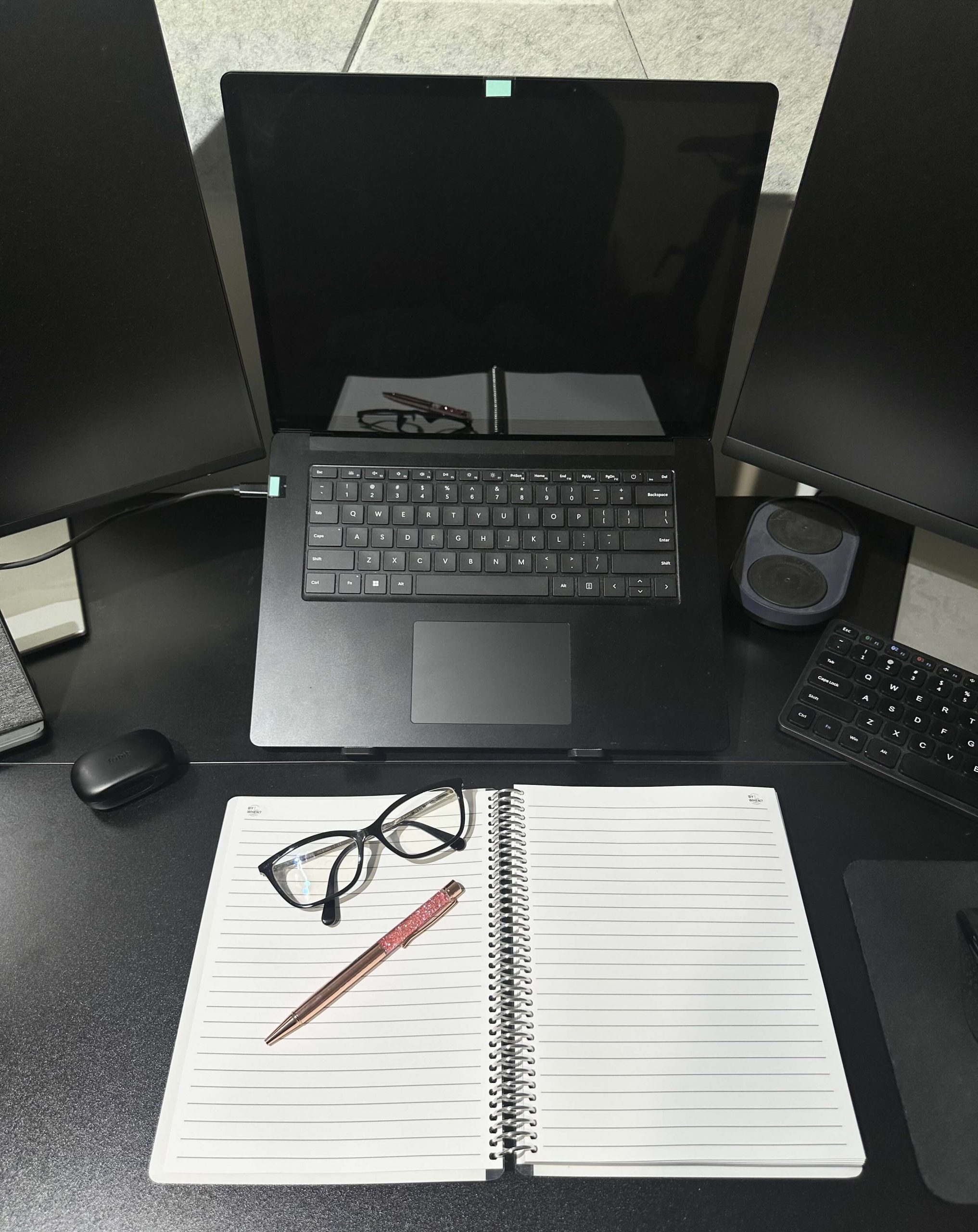 An open notebook, pen, and glasses on a desk with a laptop, monitors, and other computer accessories.