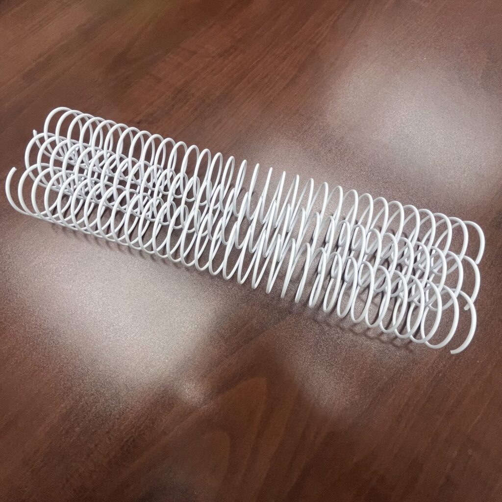 A 1+1/2" x12" #2.5 White Spiral Wire (1 Piece) rests on a brown wooden surface.