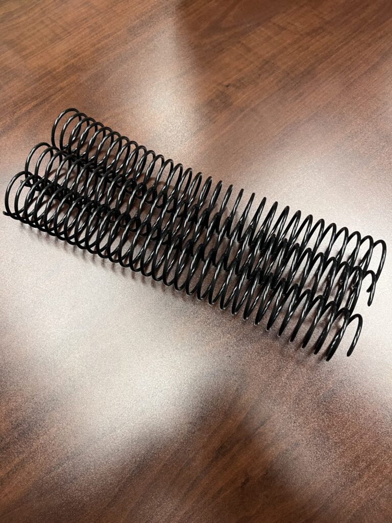 A 1+1/2" x12" #2.5 Black Spiral Plastic binding coil lies on a polished wooden surface.