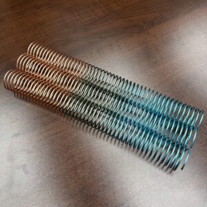 A 1+1/4" x12" #4 Paradise Solid Spiral, featuring copper, green, and blue gradient colors, lies parallel to two identical coils on a brown wooden surface.