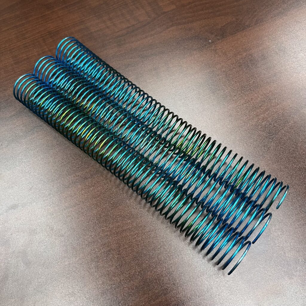 A 1+1/4” x12” #4 Blue Burst Rainbow Spiral coil spring is displayed on a brown wooden surface.