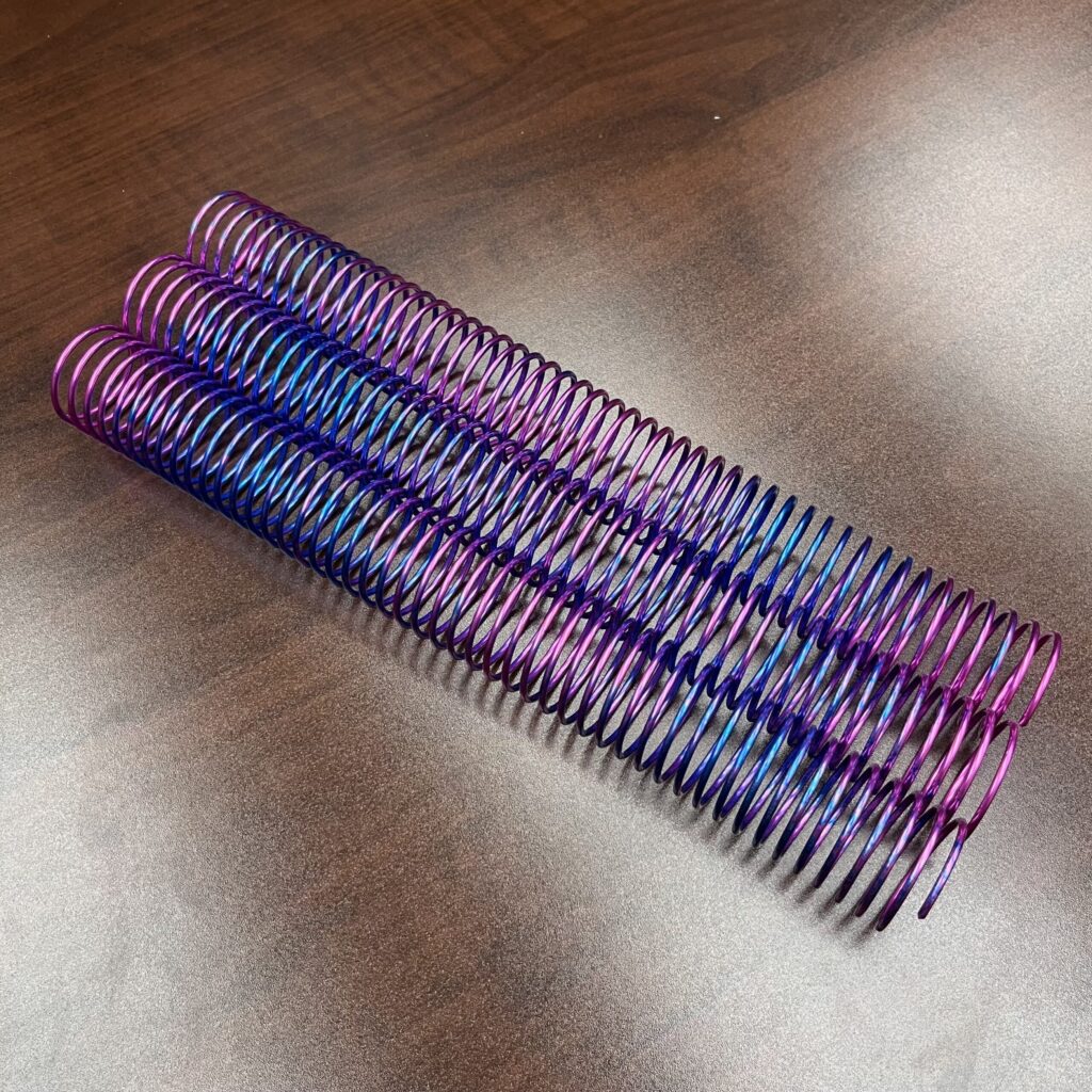 A 1+1/4” x12” #4 Purple Burst Rainbow Spiral binder coil rests on a wooden surface.