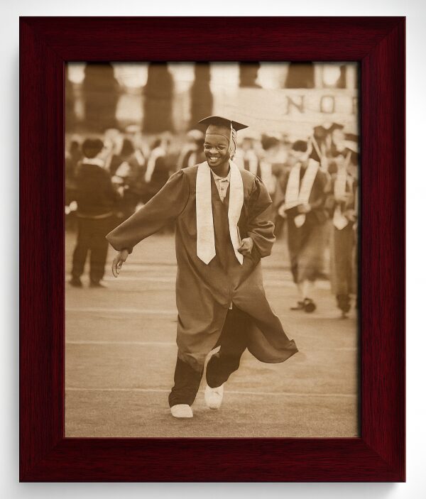 Sepia photo of a smiling graduate in cap and gown walking on a field, framed in dark wood.
