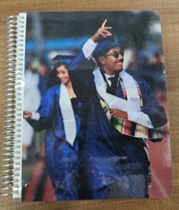 Two graduates in blue gowns celebrating, one raising hand and smiling, on the cover of a spiral notebook.