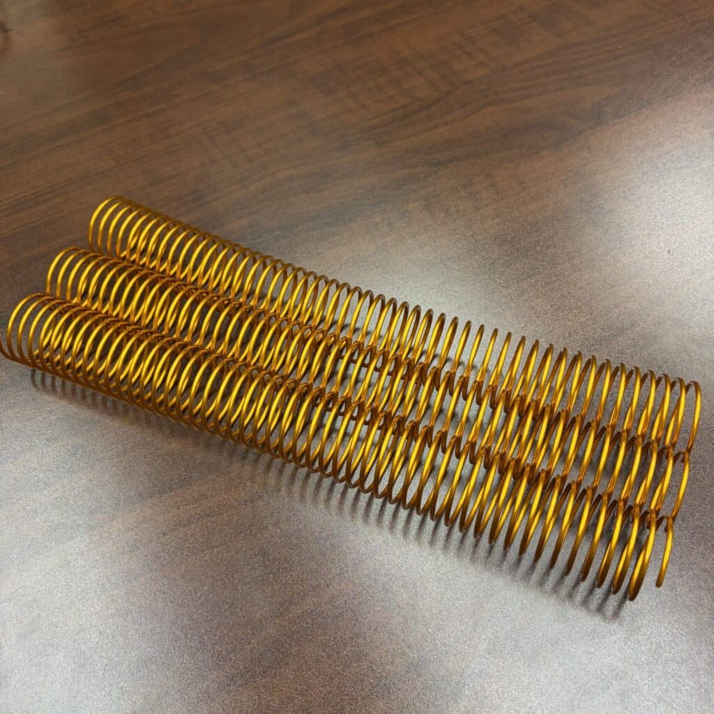 A single orange spiral aluminum piece, measuring 1+1/4″ x 12″ and featuring a #4 multicolor finish, lies on a brown wooden surface.