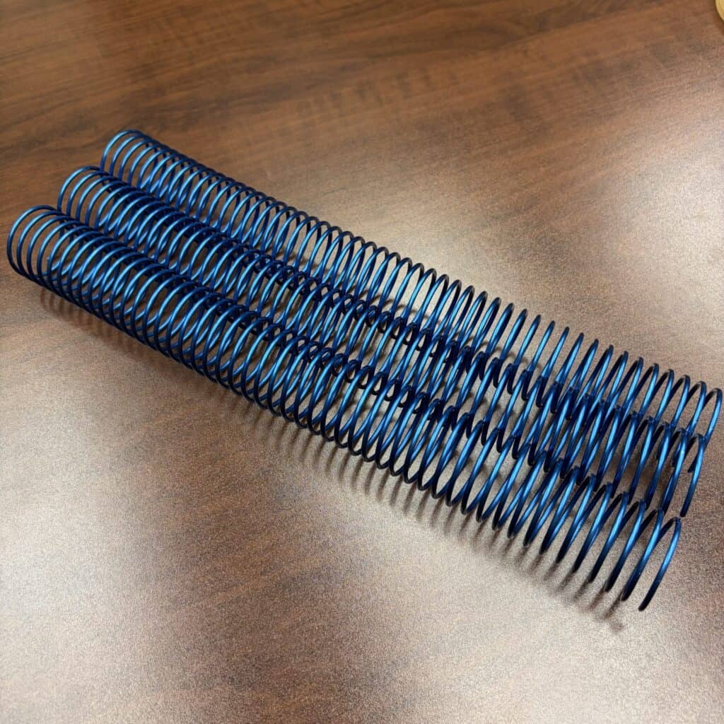 Product Rewrite: A single blue #4 solid spiral aluminum coil, measuring 1+1/4″ x 12″, lies parallel to the others on a brown wooden surface.