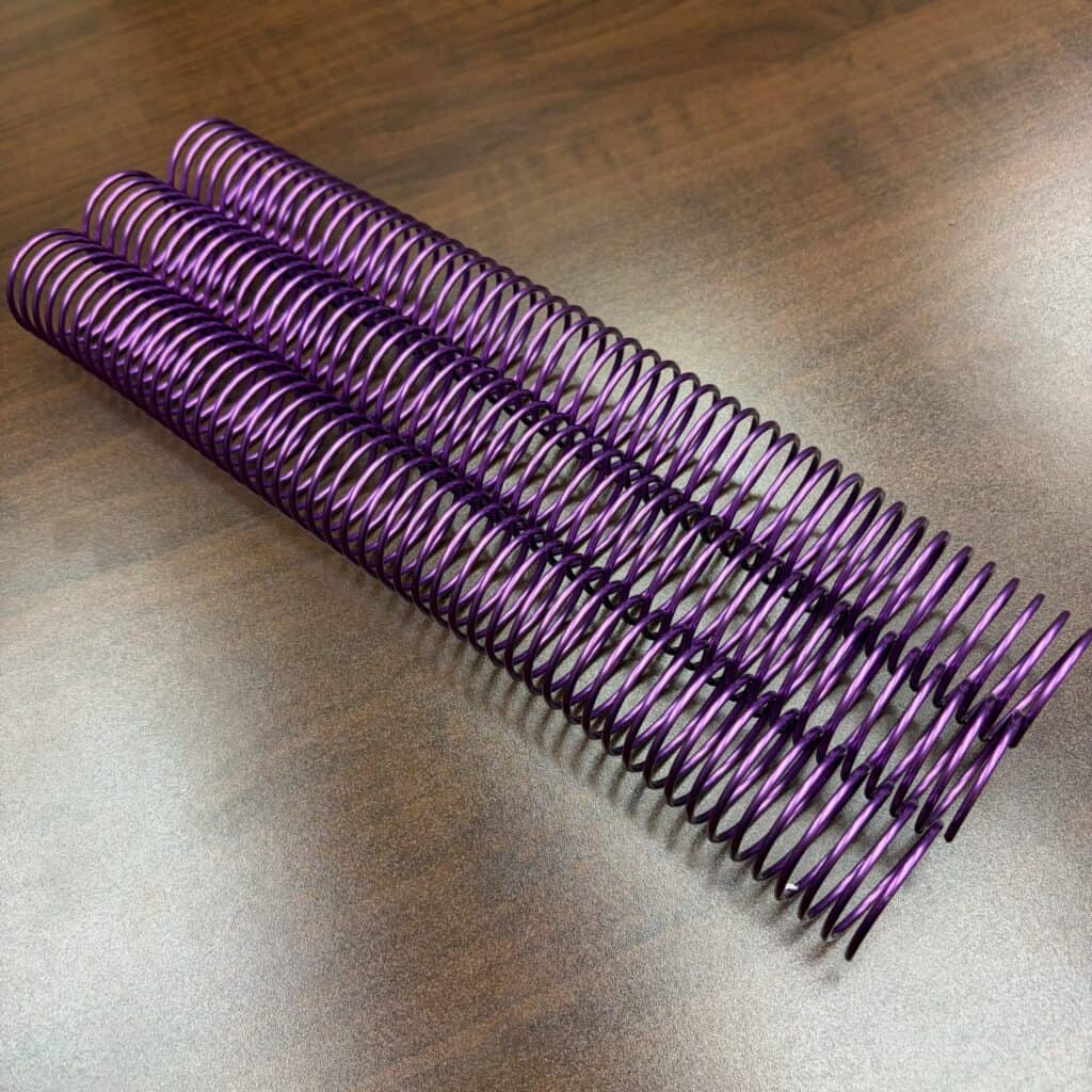 Three 1+1/4″ x 12″ #4 Violet Solid Spiral Aluminum binding spines are placed side by side on a wooden surface.