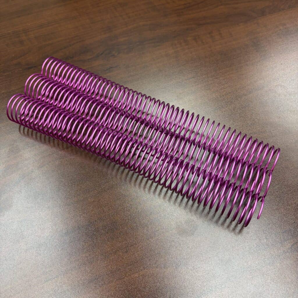 Three 1+1/4″ x12″ #4 Neon Pink Solid Spiral Aluminum coil bindings are placed side by side on a brown wooden surface.