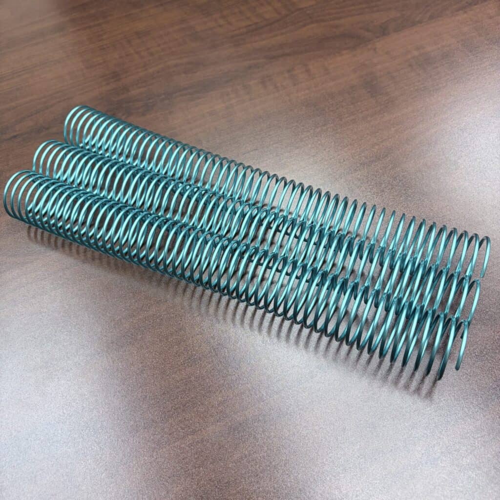 Three 1+1/4″ x12″ #4 Teal Solid Spiral Aluminum pieces are aligned parallel on a brown wooden surface.