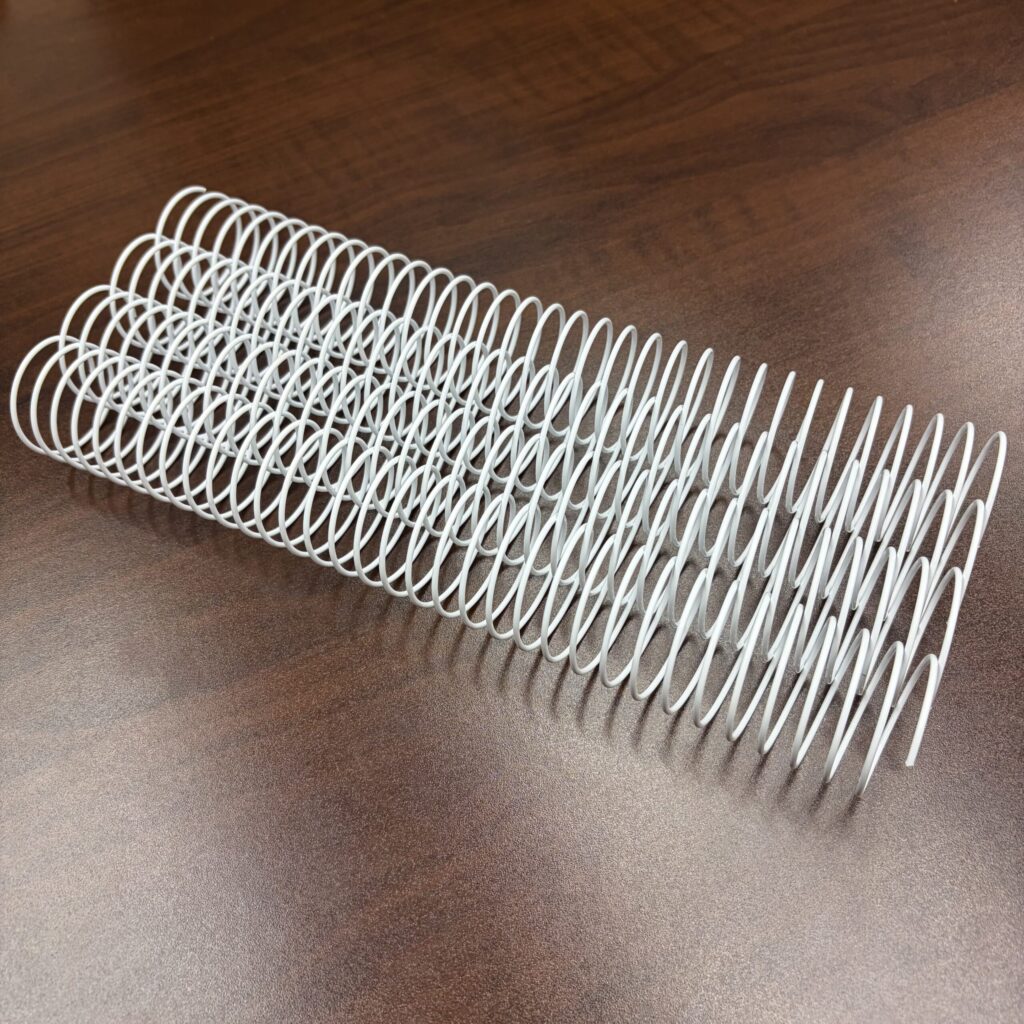 A 1+5/16″ x 10″ #.312 White Wire (1 Piece) lies on a wooden surface, commonly used for binding notebooks or documents.
