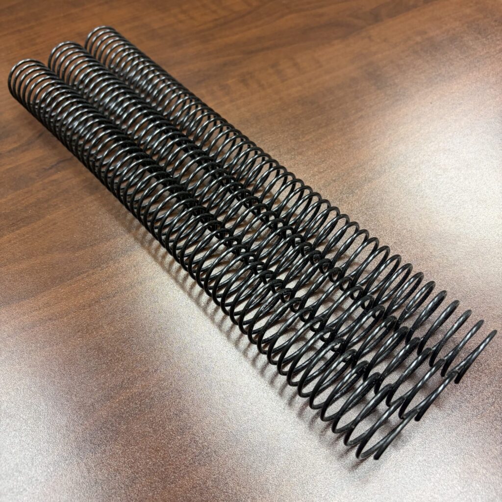 Three 1″ x12″ #4 Black Spiral Wire coils are lined up parallel to each other on a wooden surface.