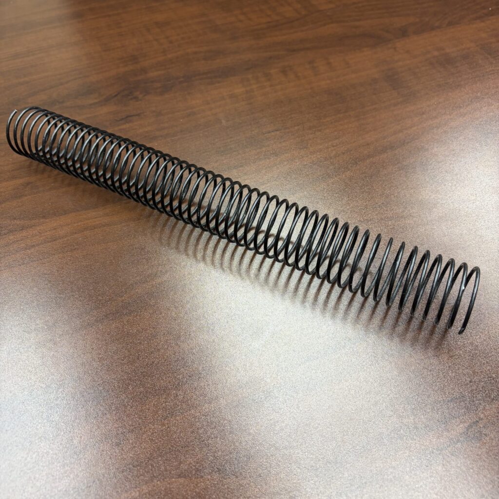 A 1+1/4″ x12″ #4 .080 black spiral wire is positioned diagonally on a wooden surface.