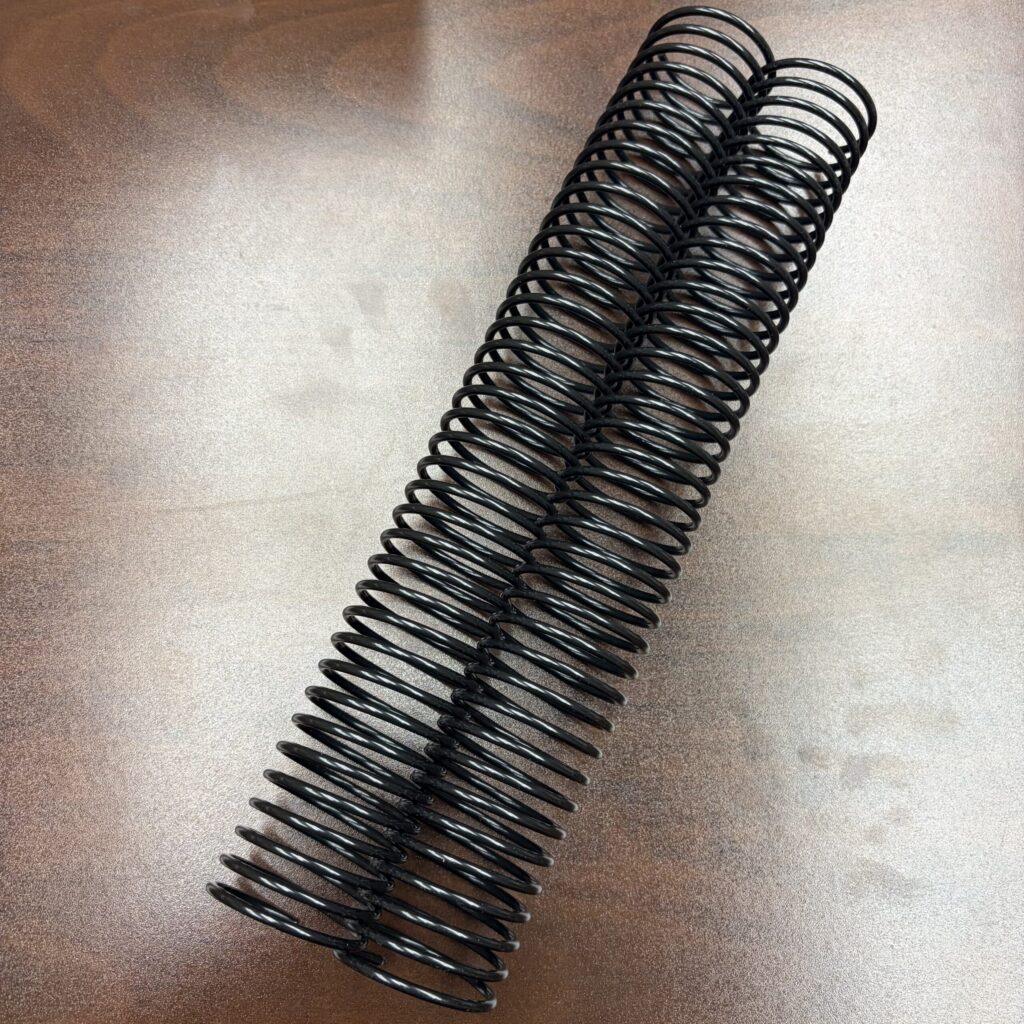 Two 1+1/2″ x12″ #3wo black spiral plastic coils (set of 2) are lying side by side on a brown wooden surface.